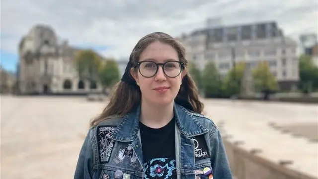 A young person with glasses and a denim jacket decorated with multiple patches and pins stands in the middle of a public square. There does not appear to be other people nearby.