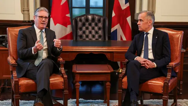 Sir Keir Starmer and Mark Carney sat on opposite sides of a table. There are Canadian and Union Jack flags hung behind the,.
