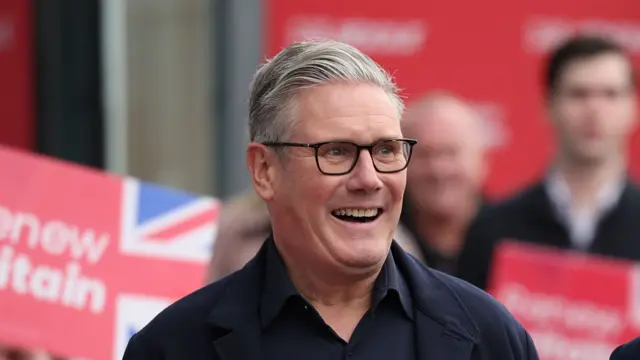 Starmer smiling. In the background is a blurred crowd as he arrives in Liverpool for the party conference. They are holding red Labour signs