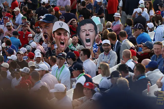 An image of fans holding up masks of Jon Rahm, Tommy Fleetwood and Rory McIlroy