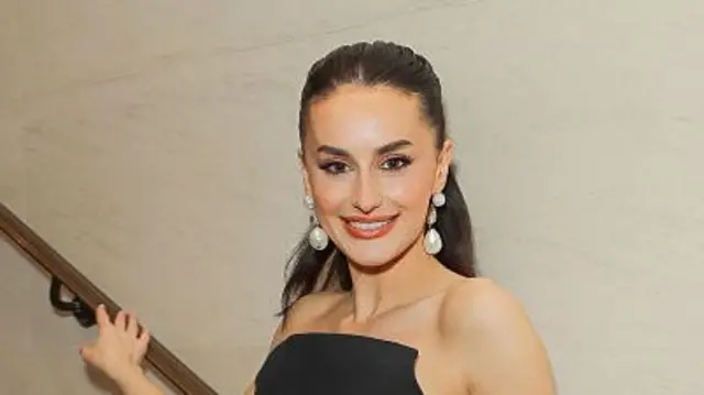 Davies smiling wearing a black cocktail strapless dress. She is posing on a staircase, with the wall behind her