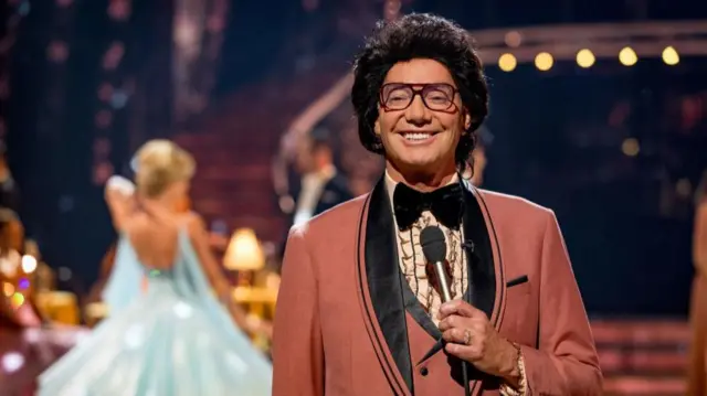 A man in a pink tuxedo and black wig stands holding a microphone on the set of Strictly.