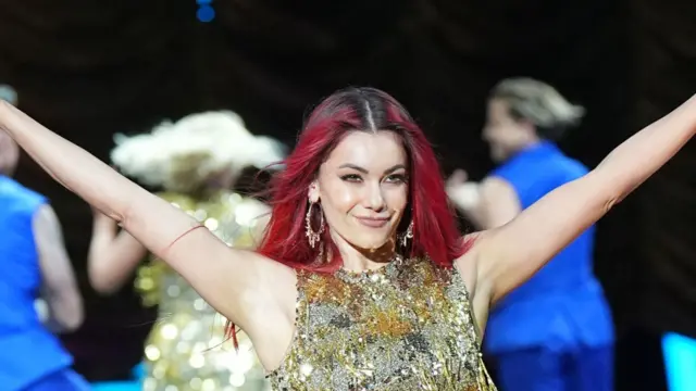 Dianne Buswell is dancing with her arms thrown out into the air, her hands out of shot. She is wearing is sparkly silver/gold sleeveless dress. Her hair - which is dark red and black at the roots - is open. Behind her are three blurred figures dancing - a blonde woman wearing the same dress, and two men wearing identical turquoise waistcoats and trousers.