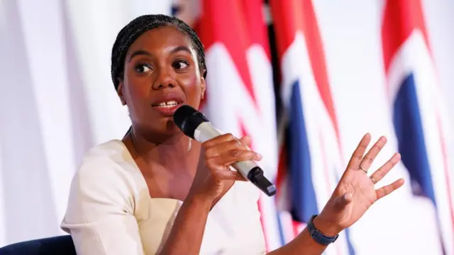 Kemi Badenoch gestures as she speaks into a microphone in front of a group of UK flags.