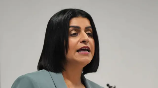 Mahmood talking into a mic that is just partially visible at the bottom of the image. She is wearing a light blue blazer, only visible from her shoulders up. She has straight black hair styled into a bob. The background is a beige wall
