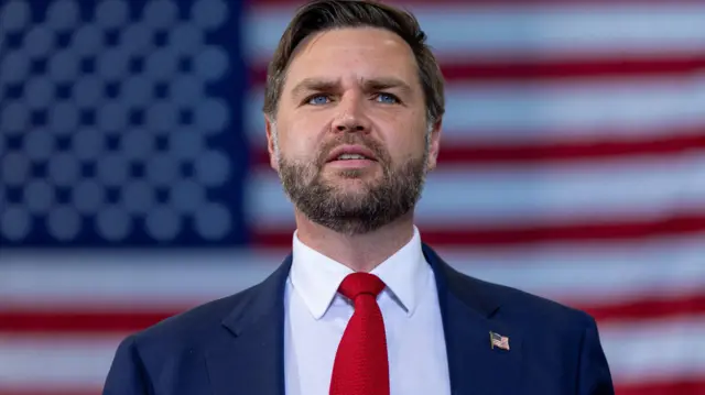 JD Vance stands in front to the US flag in a blue blazer, white shirt and red knitted tie.