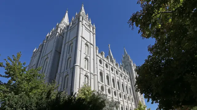 A large white religious building rises from green leafy shrubbery, with ornate turrets that reach into a blue sky.