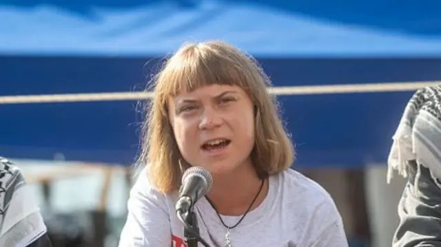 Greta in the centre with a mic in front of her. There is a person on either side of her, but they are out of frame. They are outdoors