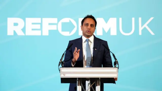 Head of policy Zia Yusuf speaking at a lectern in front of a banner reading 'Reform UK'.