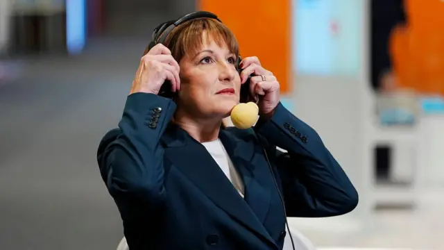Chancellor Rachel Reeves wearing a blazer holding mic headset to her ears