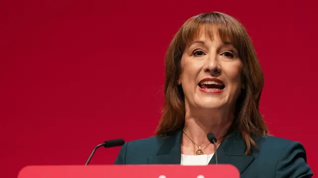 Close up of Rachel Reeves in a black blazer and white shirt as she delivers a speech at Labour conference. Behind her is wall in Labour red