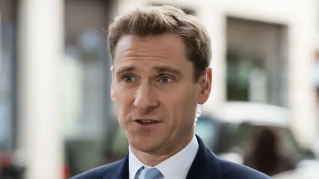 Close up of shadow Home Secretary Chris Philp in a dark blue suit, white shirt and light blue tie speaking with a clear headpiece in his left ear