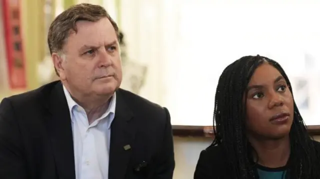 A man in a black suit sits next to a woman in a blue blouse and black jacket