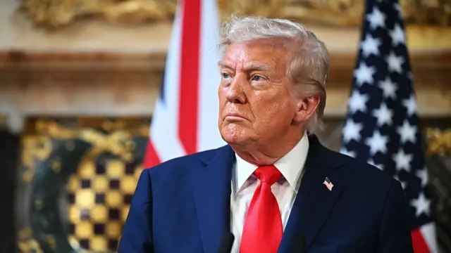 Donald Trump in a blue blazer, white shirt and red tie. He is facing to the left and behind him is a blurred outline of the US flag.