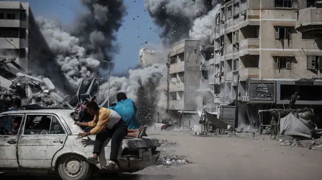 Two men hide behind a damaged car as smoke and debris flies from building behind them in Gaza City.