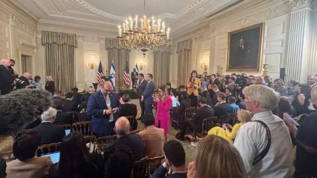 A crowd of people inside a grand room in the White House, a large framed painting of Abraham Lincoln on the right hand side of the image.
