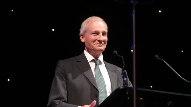 A man in a grey suit and pale green tie stands at a podium