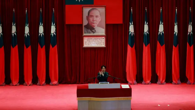 Taiwanese President William Lai speaks at a red podium. Behind him several Taiwanese flags are draped in the auditorium. In the centre, is a photo of Sun Yat-sen