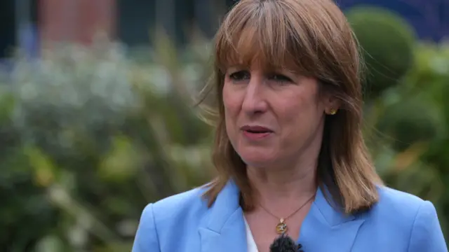 Reeves outside in front of green bushes, wearing a blue blazer