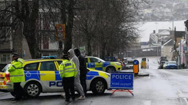 A street with two police cars parked and police officers speaking to two people
