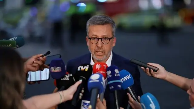 A man with glasses and grey hair speaks to a clump of reporters