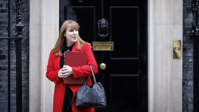 Woman in red coat, holding leather handbag and red binder, stands outside in front of a black door