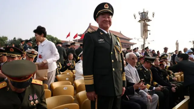 An old man in a military uniform beams at the camera, standing among other seated veterans