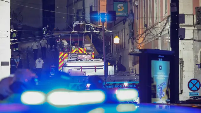 Wreckage of the funicular seen through the flashing lights of a police car. Firetrucks and first responders surround the area