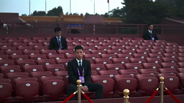 Men in suits and purple lanyards sit on empty seats outdoors