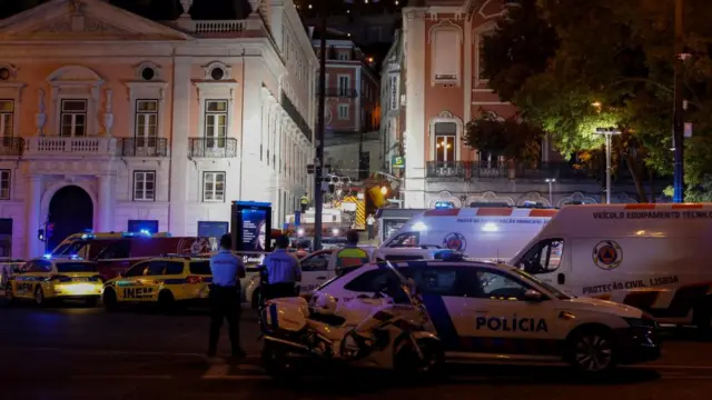 The scene of the crash in the evening, the scene illuminated by blue lights of emergency response vehicles that surround the narrow, steep cobblestone street where the wreckage lies