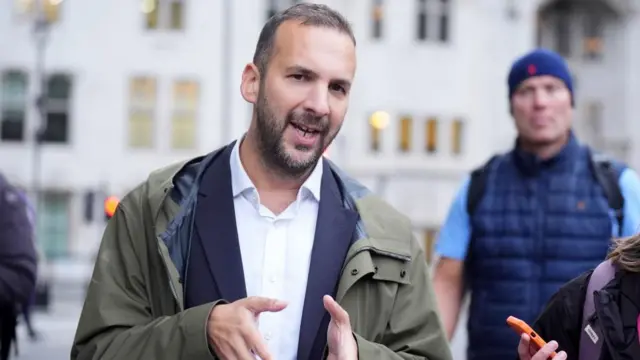 Zack Polasnki, standing outside and wearing a khaki overcoat, speaks, gesturing with his hands