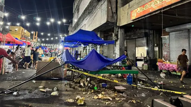 A collapsed blue gazebo is shown in a busy market street, surrounded by debris and taped off with yellow caution tape.