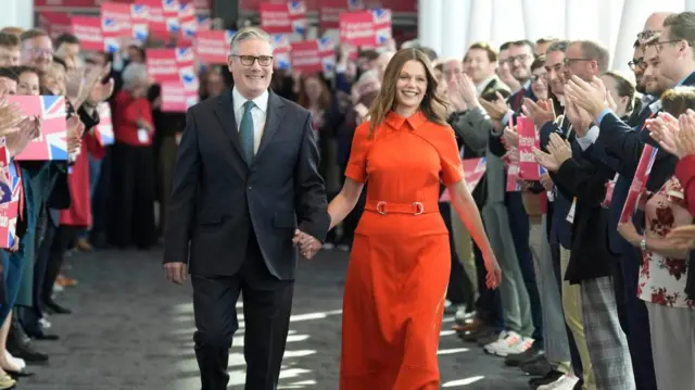 Starmer and Victoria walking up a corridor which has people lined up on either side. Everyone is smiling while the people clap them on as they walk, some holding posters in support of Labour