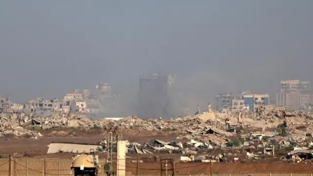 The sky is thick with smoke while piles of rubble lie on the ground. Some buildings can be seen standing in the background