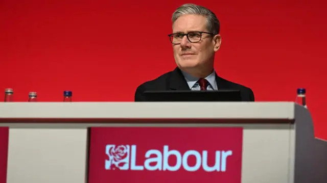 Starmer sits behind a desk with labour branding, in front of a bright red backdrop