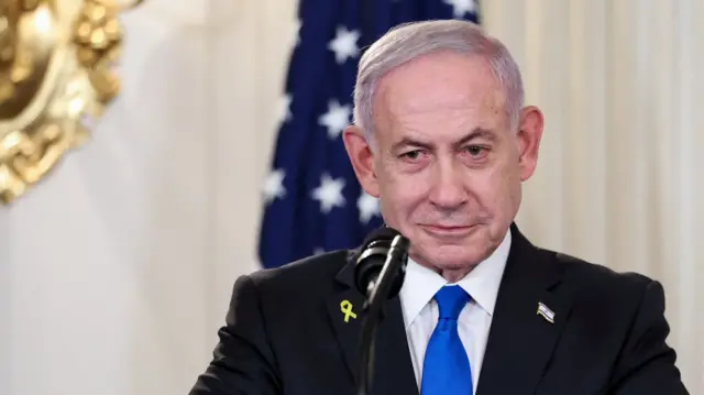 Close up of Benjamin Netanyahu during a joint press conference at the White House. He's in a dark suit with a blue tie, he's wearing a yellow ribbon pin on his right lapel and an Israeli flag pin on his left one