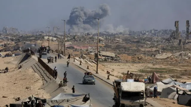 Smoke rises during an Israeli military operation as displaced Palestinians fleeing northern Gaza, cars and vans on the road as people walk nearby.