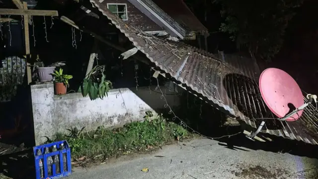 A corrugated iron roof with a red satellite dish is shown collapsed over a white concrete wall
