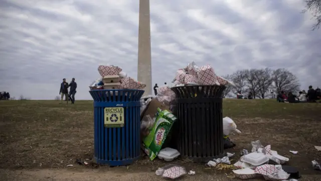 Two full bins overflowing with cardboard food boxes on a grey wintery day. They are in front of the large Washington Monument, which has people walking around on the grass between it and the bins