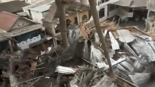 A screengrab of a video showing a collapsed roof filmed from above, corrugated iron, concrete, steel bars and other construction materials are covering the ground.