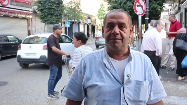 Sadeq Kamil stands in front of the camera as people walk behind him