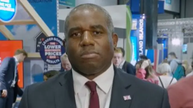 Headshot of Lammy who stands in a busy hall in front of stalls with lots of people milling around