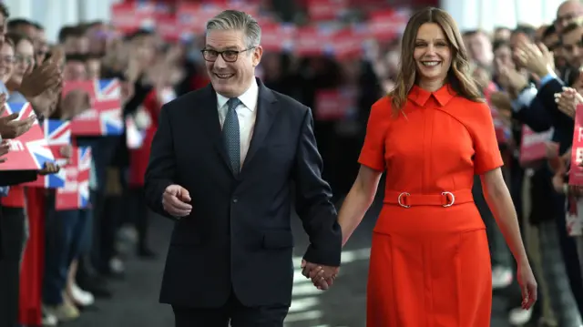 Starmer points and smiles as he walks down a corridor