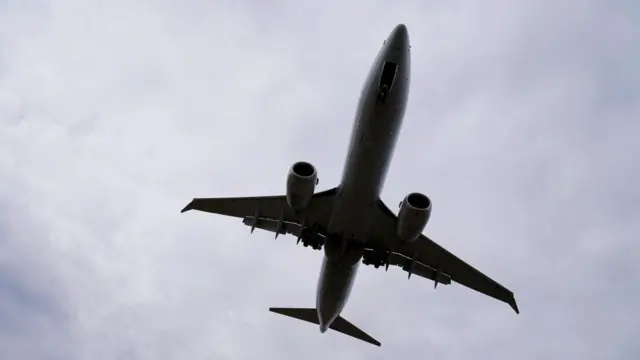 A stock image of a plane flying above the camera. It's dark and in a shadow against a white cloudy sky.