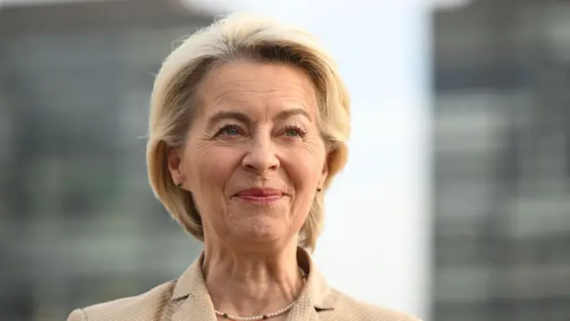 Von der Leyen in a beige jacket in front of a grey blurred background.