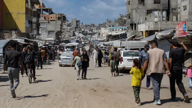 A group of people walking down a street in southern Gaza, cars, buildings and small stalls with canopies over them line the street.