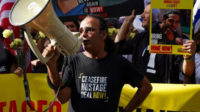 A man with a black t-shirt and a megaphone, the t-shirt reads "ceasefire, hostage deal now"