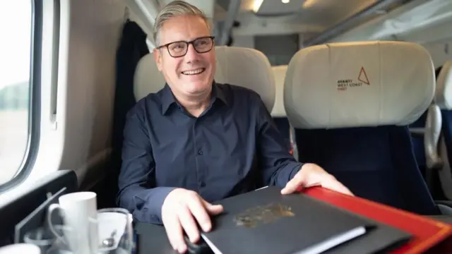 Keir Starmer in a dark shirt, smiling as he holds a pile of folders. He is sitting in a window seat in an Avanti West Coast first class train carriage on his way to Liverpool