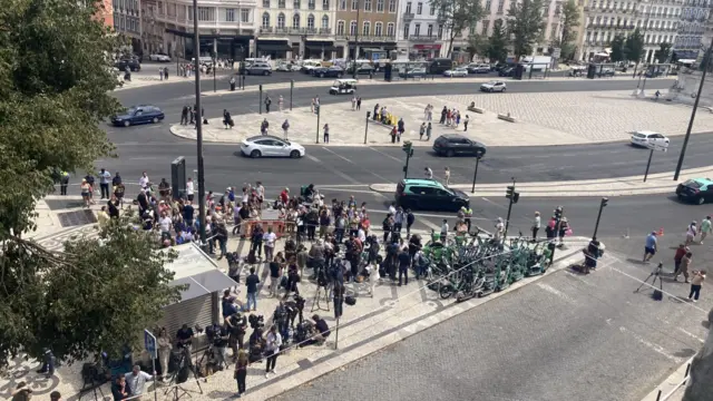 A crowd of journalists with cameras stand outside a cordon, e-bikes crowded on pavement.