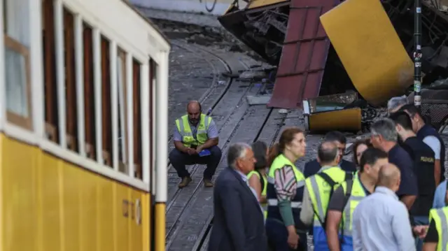 People stand near a crashed funicular carriage that is yellow and white. They wear high vis jackets. Another carriage is on the tracks in the foreground of the image.